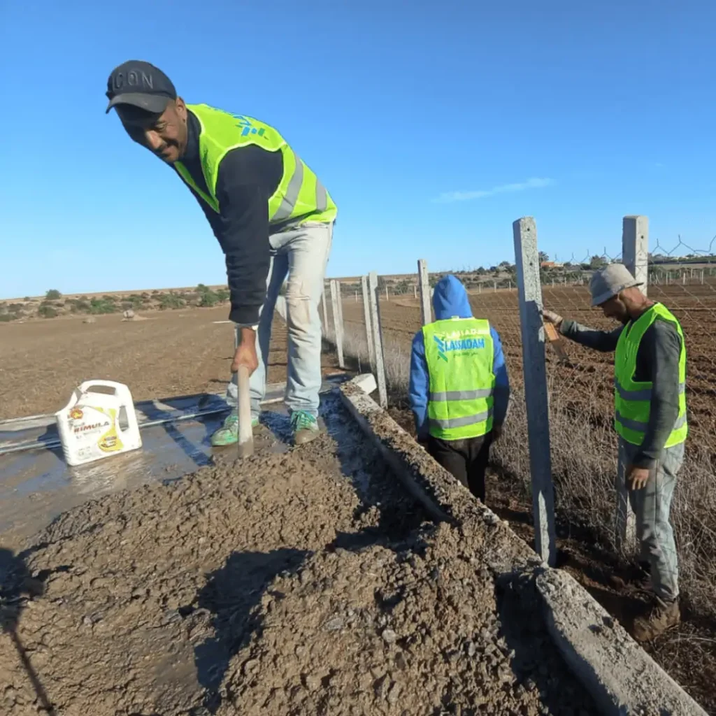 Ouvriers préparant et coulant du béton pour les fondations et le scellage des poteaux d'une clôture de terrain.